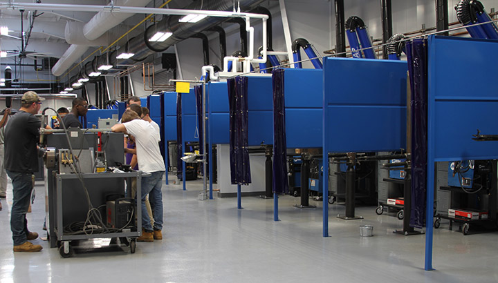 Row of weld booths in a lab featuring Miller power sources, along with students working together 