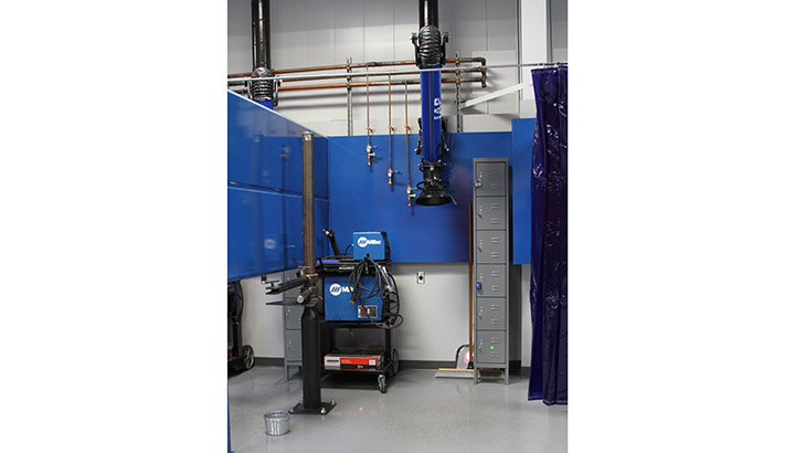 Inside of weld booth feature a Miller power source and wire feeder, along with a fume extraction arm and fixture for weld coupons