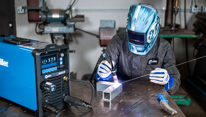 Operator sits at a table in a shop and TIG welds on a part with a Dynasty TIG welder
